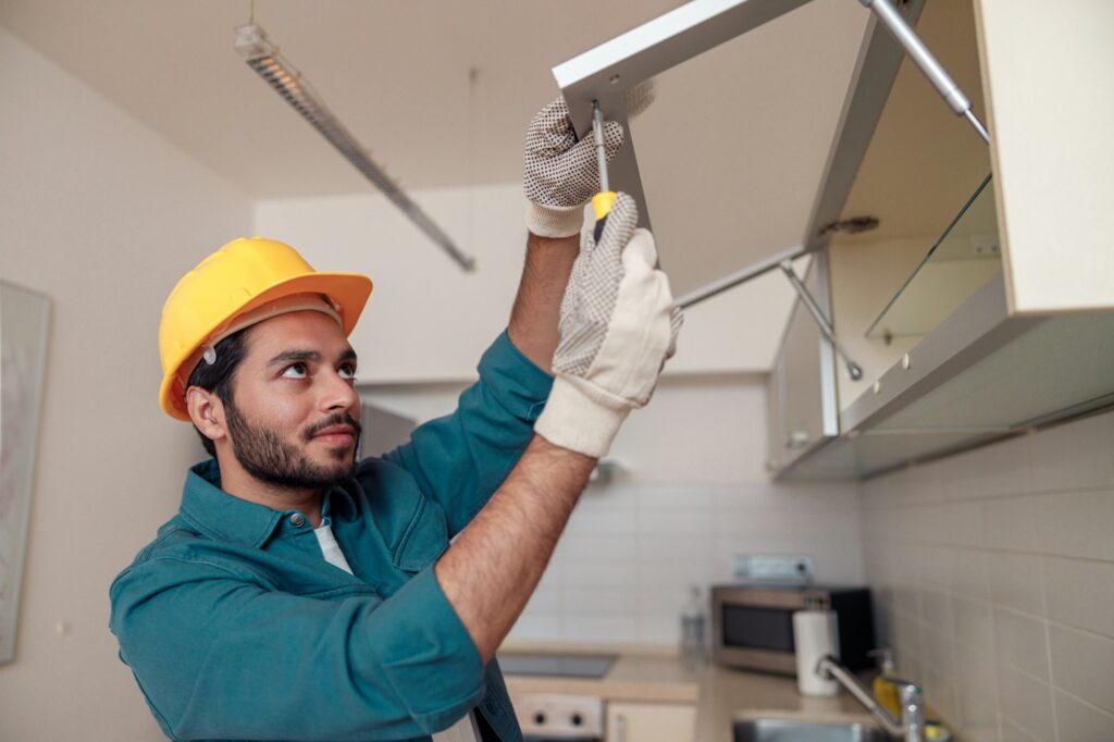 Focused professional worker in uniform is assembling furniture on kitchen. Repair concept